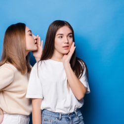 young-woman-whispers-her-mate-bad-news-isolated-blue-wall